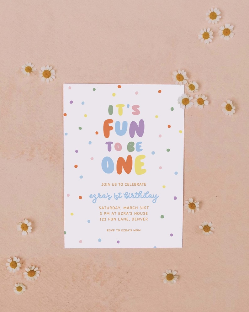 May include: A white invitation card with the text "IT'S FUN TO BE ONE" in colorful letters and confetti dots. The card announces Ezra's 1st birthday party, including the date, time, and location. Small white daisies are scattered around the card.