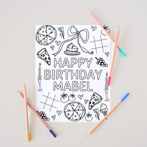May include: A coloring book page with the text "HAPPY BIRTHDAY MABEL" surrounded by line drawings of pizza, cake, hearts, and other celebratory images. Several colored pencils are scattered around the page.