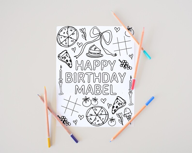 May include: A coloring book page with the text "HAPPY BIRTHDAY MABEL" surrounded by line drawings of pizza, cake, hearts, and other celebratory images. Several colored pencils are scattered around the page.
