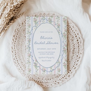 May include: A bridal shower invitation with the text "Olivia's Bridal Shower" in an oval frame, surrounded by floral and striped patterns. The invitation is on a cream-colored crocheted doily, with dried wheat stalks in the background.