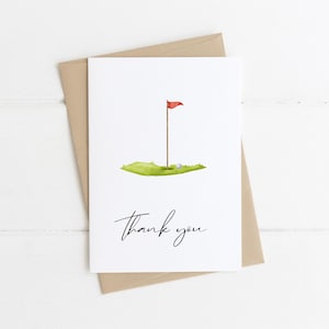 Könnte beinhalten: Weiße Grußkarte mit einer Aquarellillustration eines Golfgrüns, einer Fahne und zwei Golfbällen. Die Worte "thank you" sind in Kursivschrift geschrieben. Die Karte liegt auf einem braunen Umschlag.