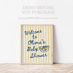 May include: A framed baby shower sign with a yellow and white striped background. The sign reads "Welcome to Olivia's Baby Shower" in navy blue script, with a lemon illustration. The sign is in a light wood frame.