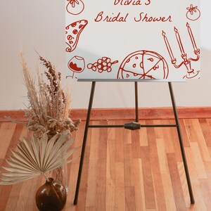 May include: A white easel with a red hand-drawn design of pizza, wine glasses, grapes, and candles. The text "Olivia's Bridal Shower" is written in red on the easel.