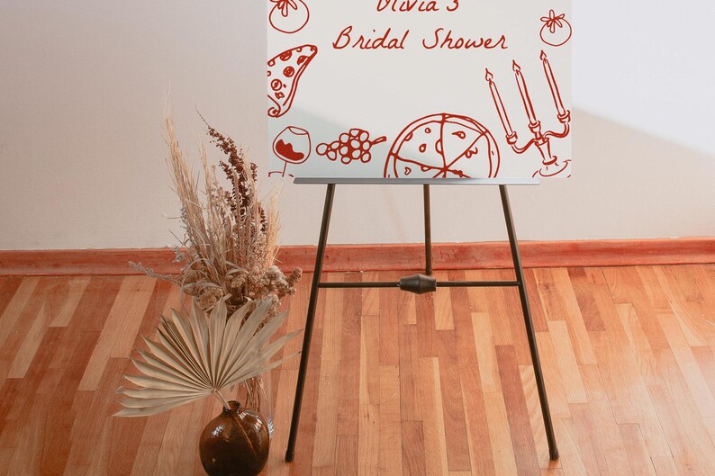 May include: A white easel with a red hand-drawn design of pizza, wine glasses, grapes, and candles. The text "Olivia's Bridal Shower" is written in red on the easel.