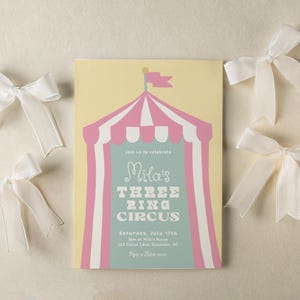May include: A circus-themed invitation with a pink and white striped tent design, the text "Mila's THREE RING CIRCUS", and white satin bows. The invitation is on a light yellow background.