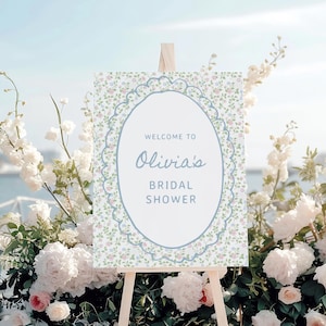 May include: A welcome sign for Olivia's bridal shower, displayed on an easel. The sign features a floral design with a blue lace-like border and the text "Welcome to Olivia's Bridal Shower". The sign is surrounded by white and pink flowers.