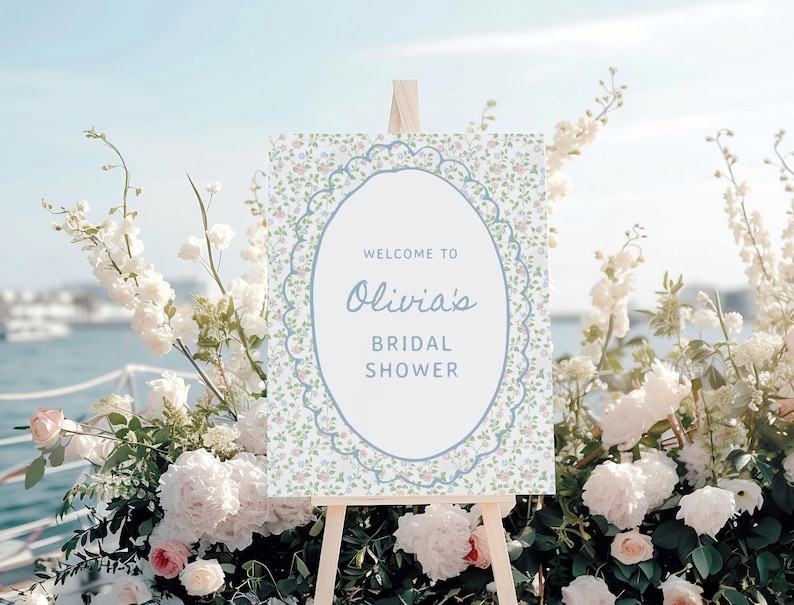 May include: A welcome sign for Olivia's bridal shower, displayed on an easel. The sign features a floral design with a blue lace-like border and the text "Welcome to Olivia's Bridal Shower". The sign is surrounded by white and pink flowers.