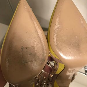 May include: A pair of yellow high-heeled shoes with gold studs. The shoes have a pointed toe and a thin strap across the foot. The soles of the shoes are beige.