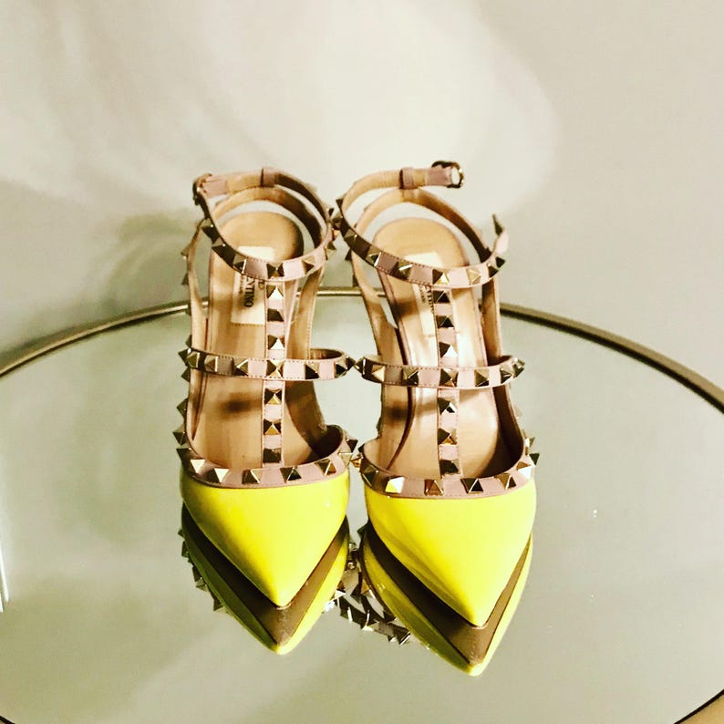 May include: A pair of yellow high-heeled shoes with gold studs and straps. The shoes are on a mirrored surface.