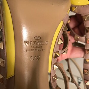 May include: Close-up of the bottom of a yellow and beige high-heeled shoe with gold studs. The shoe is labeled "VALENTINO GARAVANI MADE IN ITALY" and "37 1/2".