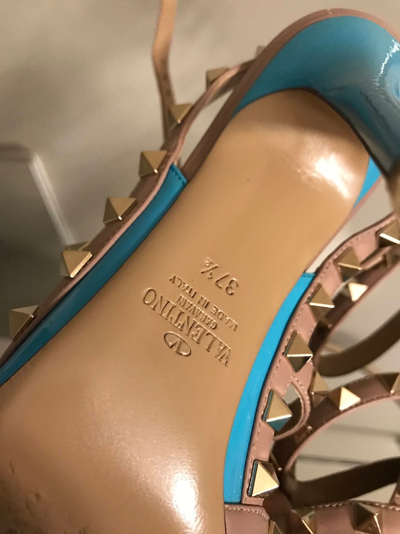 Puede incluir: Primer plano de la parte inferior de un zapato de tac&oacute;n alto rosa claro y azul con tachuelas doradas. El zapato est&aacute; etiquetado como "VALENTINO GARAVANI MADE IN ITALY 37 1/2".