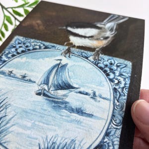 May include: A watercolor painting on wood featuring a sailboat scene within a blue and white decorative border. A small bird perches on the edge of the painting. The artwork has a rustic, handmade aesthetic.