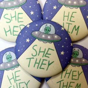 May include: Several round badges with an alien design. Each badge shows a green alien in a flying saucer, with the phrases "SHE HER", "HE HIM", "SHE THEY", and "THEY THEM" in green text. The background is blue with white stars.