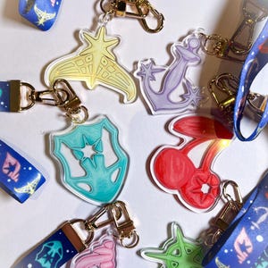Puede incluir: Una colección de llaveros acrílicos con cierres dorados y cordones azules. Los llaveros presentan varios diseños, incluyendo una estrella amarilla, un ancla púrpura, un escudo turquesa, cerezas rojas, una concha rosa y una llave verde.