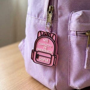 Pode incluir: Uma mochila de lona roxo-claro com um porta-chaves rosa em forma de mochila preso ao fecho. O porta-chaves apresenta um contorno preto e texto branco que diz CONTACTO DE EMERGENCIA e SOFIA, com um pequeno ícone de sinal no topo.