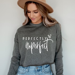 May include: A grey cropped sweatshirt with the text "PERFECTLY imperfect" in white lettering with a small heart symbol. The model is wearing the sweatshirt with blue jeans and a brown hat.