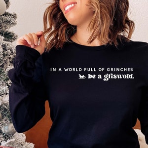 Op de afbeelding: Een vrouw draagt een zwart shirt met lange mouwen met de tekst "In een wereld vol Grinches, wees een Griswold." Ze draagt een rode en zwarte geruite pyjamabroek en zit voor een met sneeuw bedekte kerstboom.