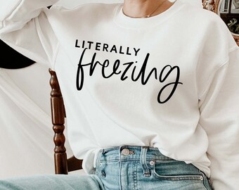 以下が含まれることがあります： 黒い文字で「LITERALLY freezing」と書かれた白いスウェットシャツ。スウェットシャツは、青いジーンズを履いた人が着用しています。