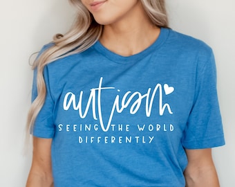 Op de afbeelding: Een blauw t-shirt met witte tekst die "autisme de wereld anders zien" zegt met een klein wit hartje na het woord "autisme".