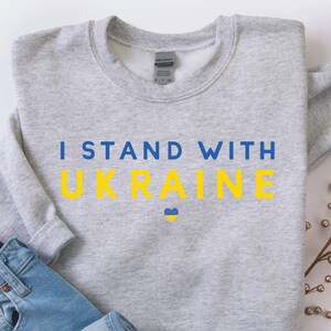 May include: A gray sweatshirt with the text "I Stand With Ukraine" in blue and yellow letters. A small yellow and blue heart is below the text.