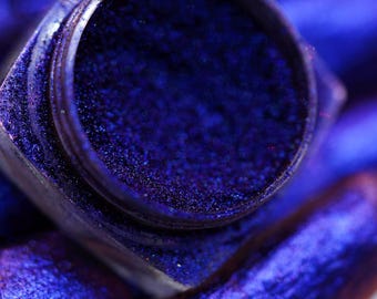May include: A small, square container filled with a deep blue iridescent glitter. The glitter is very fine and has a shimmery finish.