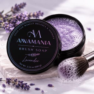 Op de afbeelding: Een zwarte ronde houder met AWAMANIA penseelzeep met lavendelgeur. Het deksel bevat de merknaam, de tekst SOAP FOR MAKEUP BRUSHES & SPONGES en 100 g. Een make-upkwast met zeepbellen ligt naast de geopende pot met zeep.