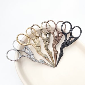 May include: A set of six small, decorative scissors with bird-shaped handles. The scissors are arranged in a circle on a white surface. The handles are in various metallic finishes, including gold, silver, bronze, and black.