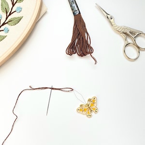 Pode incluir: Materiais de bordado em fundo branco. Inclui um bastidor de madeira com design floral, linha marrom, uma agulha, tesouras douradas e um pingente em forma de borboleta com detalhes amarelos. Para projetos de artesanato.