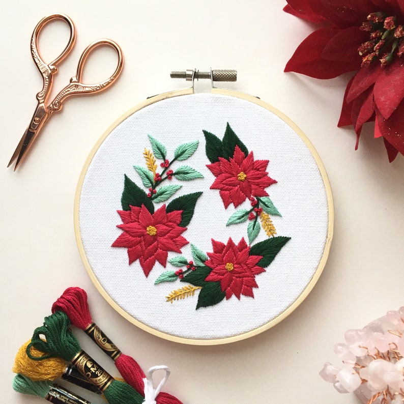 May include: A white embroidery hoop with a red and green floral design. The design features poinsettia flowers and green leaves.