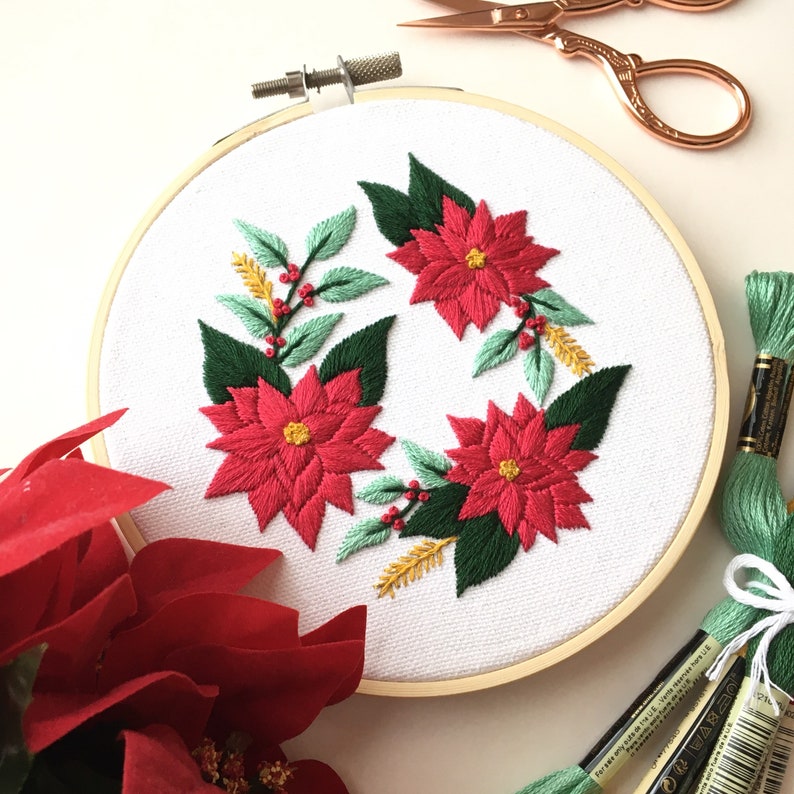May include: Embroidered hoop art featuring a wreath of poinsettia flowers with green leaves and red berries. The flowers are red with yellow centers and the leaves are a deep green. The hoop is made of wood and has a white background.