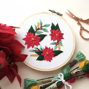 May include: A white embroidery hoop with a red and green poinsettia floral design. The hoop is surrounded by spools of embroidery floss in various colors.