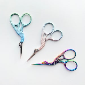 May include: Three pairs of decorative stork scissors in various colors. One pair is blue and green, another is pink and silver, and the third is a rainbow of colors. The scissors have a bird-like design.
