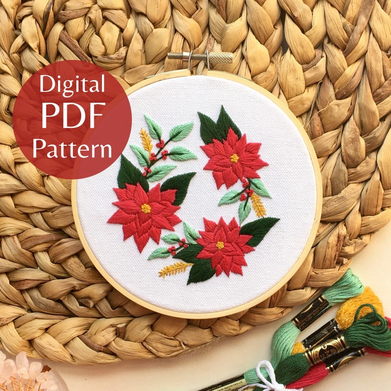 May include: A digital PDF embroidery pattern featuring a wreath of poinsettia flowers, leaves, and berries. The pattern is displayed in a round embroidery hoop on a woven wicker surface.