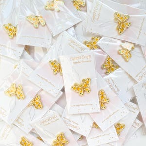 Pode incluir: Uma cole&ccedil;&atilde;o de passa-fios em forma de borboleta. Cada alfinete de borboleta dourado apresenta detalhes em esmalte amarelo e verde. Os alfinetes s&atilde;o embalados em cart&otilde;es brancos com o texto "Eight22 Crafts Needle Threader" e um endere&ccedil;o web.