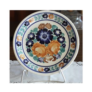 Könnte beinhalten: Ein weißer Keramikteller mit einem blauen und orangefarbenen Blumendesign. Der Teller hat einen weißen Hintergrund mit einem blauen und grünen Rand. Das Blumendesign zeigt blaue, orange und grüne Blumen.