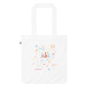 May include: White tote bag with a colourful doodle design featuring the word "hibbli" and various objects such as sandals, flowers, and a glass of wine. The text "Team with Purpose" is also included in the design.