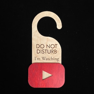May include: Wooden door hanger with the text "DO NOT DISTURB I'm Watching". The hanger has a natural wood finish with a red YouTube logo at the bottom. The background is black.