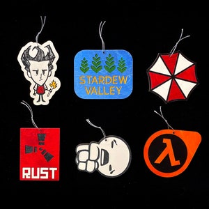 Puede incluir: Conjunto de seis adornos de madera con varios personajes de videojuegos y de la cultura popular. Los adornos están pintados en varios colores, incluyendo rojo, azul, verde, negro, blanco y naranja. Los adornos representan los siguientes personajes: Rust, Half-Life, Stardew Valley, una criatura del juego Don't Starve y el logotipo de Umbrella Corporation.