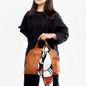 May include: A woman is holding a brown leather satchel bag with a white and orange patterned scarf hanging from the handle.