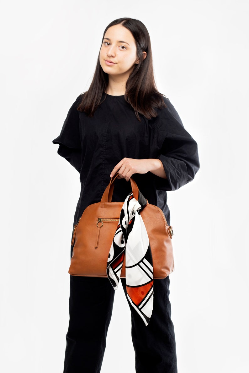 May include: A woman is holding a brown leather satchel bag with a white and orange patterned scarf hanging from the handle.