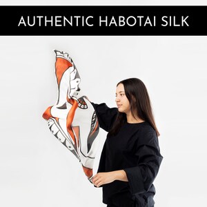May include: A person holds up a white silk scarf with a black and orange abstract design. The scarf is made of authentic Habotai silk.