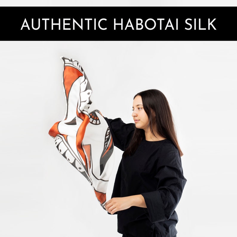 May include: A person holds up a white silk scarf with a black and orange abstract design. The scarf is made of authentic Habotai silk.