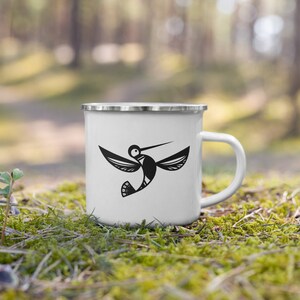 May include: White enamel mug with a black hummingbird design. The bird is depicted in flight with its wings spread. The mug has a silver rim and a white handle. The mug is sitting on a bed of green moss.