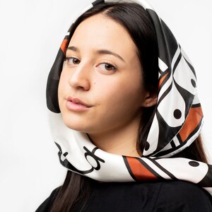 May include: A woman with long dark hair is wearing a white headscarf with a black and orange geometric pattern. The scarf is tied around her head and neck.