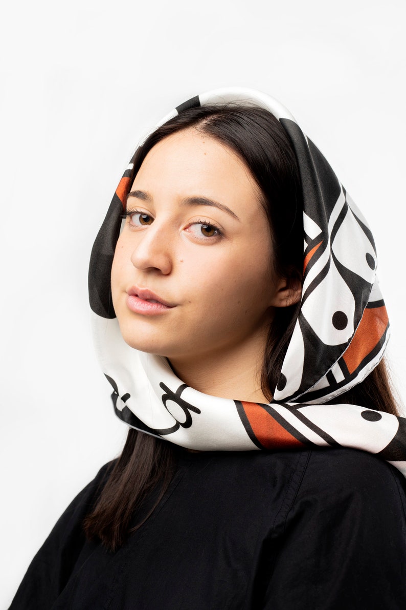 May include: A woman with long dark hair is wearing a white headscarf with a black and orange geometric pattern. The scarf is tied around her head and neck.