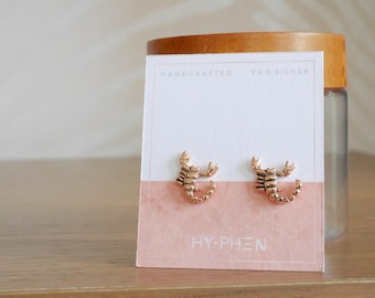 May include: A pair of gold-toned scorpion stud earrings on a white card with the text "HANDCRAFTED 925 SILVER HY-PHEN".