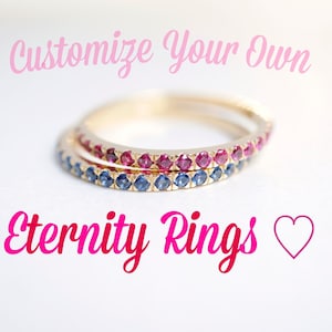Puede incluir: Dos anillos de eternidad de oro con una fila de peque&ntilde;as piedras preciosas azules y rojas. Los anillos est&aacute;n apilados uno encima del otro. El texto "Customize Your Own Eternity Rings" est&aacute; escrito en rosa sobre los anillos. Un coraz&oacute;n rosa est&aacute; debajo del texto.