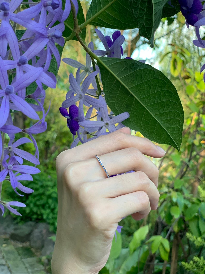 Puede incluir: Un delicado anillo de plata con una fila de peque&ntilde;as piedras azules. El anillo se lleva en una mano sobre un fondo de flores moradas.
