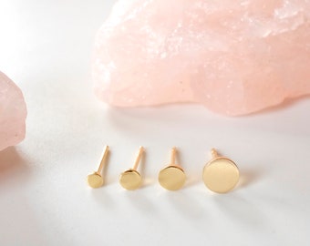 May include: Four gold-colored stud earrings of different sizes, arranged in a row on a white surface.