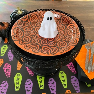 May include: A black and orange Halloween themed paper plate with a white ghost on top. The plate is on a black metal stand. The stand is on a black and green table runner with a pattern of coffins and bats.